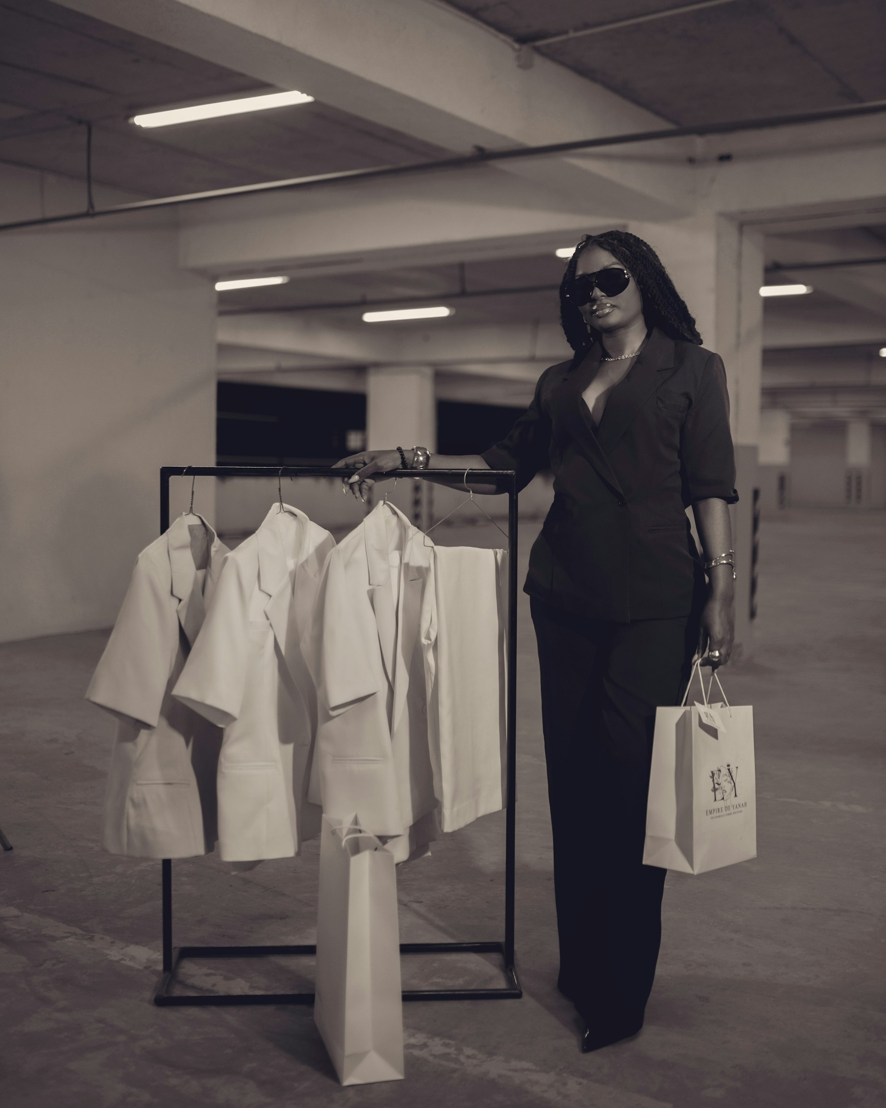 A woman in a black suit and sunglasses stands beside a clothes rail, holding a shopping bag